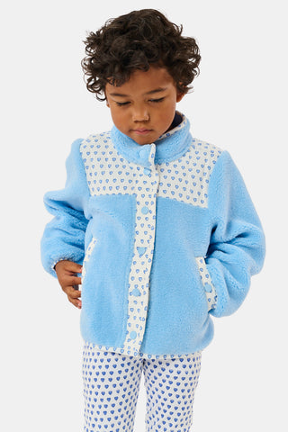 A young girl stands and smiles against a plain background, wearing the Roller Rabbit Kids Hearts Lizzie Fleece Jacket in light blue with white patterned details, paired with matching white leggings adorned with blue hearts. This cozy outerwear features soft fleece for comfort and warmth.