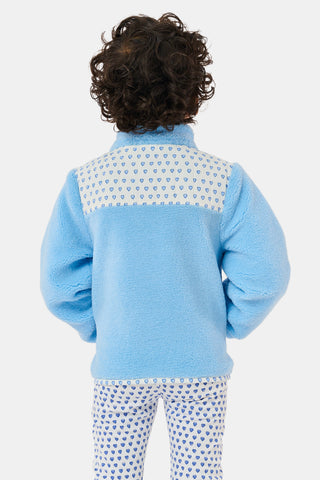 A young girl stands and smiles against a plain background, wearing the Roller Rabbit Kids Hearts Lizzie Fleece Jacket in light blue with white patterned details, paired with matching white leggings adorned with blue hearts. This cozy outerwear features soft fleece for comfort and warmth.