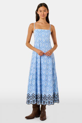 A woman wearing the Roller Rabbit Lakai Asher Dress—a light blue patterned cotton batiste dress with dark lace trim—walks on grass near a cream-colored house with green shutters, partially visible.