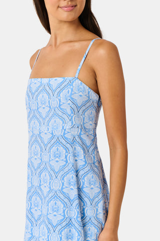 A woman wearing the Roller Rabbit Lakai Asher Dress—a light blue patterned cotton batiste dress with dark lace trim—walks on grass near a cream-colored house with green shutters, partially visible.