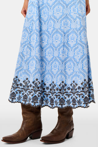A woman wearing the Roller Rabbit Lakai Asher Dress—a light blue patterned cotton batiste dress with dark lace trim—walks on grass near a cream-colored house with green shutters, partially visible.