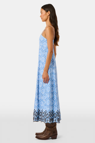 A woman wearing the Roller Rabbit Lakai Asher Dress—a light blue patterned cotton batiste dress with dark lace trim—walks on grass near a cream-colored house with green shutters, partially visible.