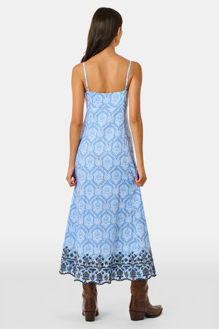 A woman wearing the Roller Rabbit Lakai Asher Dress—a light blue patterned cotton batiste dress with dark lace trim—walks on grass near a cream-colored house with green shutters, partially visible.