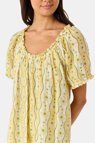 A girl wears the Roller Rabbit Kids Lillian Floral Stripe Tristan Dress, a cotton sundress with green and white stripes, floral patterns, ruffled straps, and a ruffled hem, standing with a smile and one arm relaxed at her side.