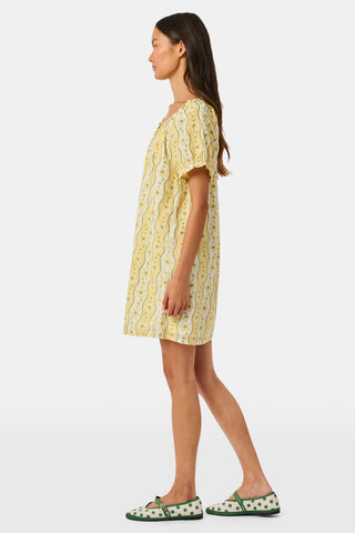 A girl wears the Roller Rabbit Kids Lillian Floral Stripe Tristan Dress, a cotton sundress with green and white stripes, floral patterns, ruffled straps, and a ruffled hem, standing with a smile and one arm relaxed at her side.