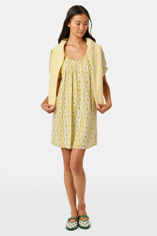 A girl wears the Roller Rabbit Kids Lillian Floral Stripe Tristan Dress, a cotton sundress with green and white stripes, floral patterns, ruffled straps, and a ruffled hem, standing with a smile and one arm relaxed at her side.