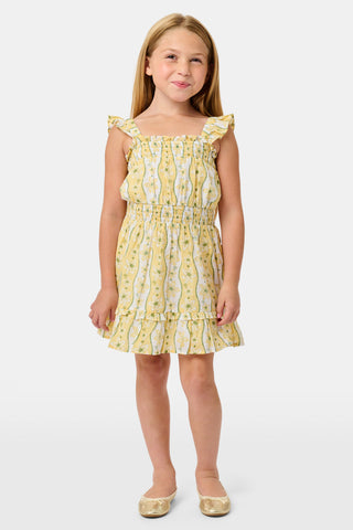 A girl wears the Roller Rabbit Kids Lillian Floral Stripe Tristan Dress, a cotton sundress with green and white stripes, floral patterns, ruffled straps, and a ruffled hem, standing with a smile and one arm relaxed at her side.