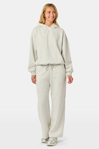 A person is wearing the Roller Rabbit Sleep Club Mimi Sweatpants in light gray, featuring wide legs and crafted from velvety soft Dream Fleece with “Sleep Club” printed in light blue on the left thigh, paired with white sneakers and a cropped white top. The background is plain white.