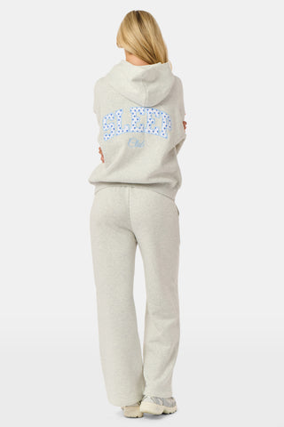 A person is wearing the Roller Rabbit Sleep Club Mimi Sweatpants in light gray, featuring wide legs and crafted from velvety soft Dream Fleece with “Sleep Club” printed in light blue on the left thigh, paired with white sneakers and a cropped white top. The background is plain white.