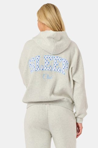 A person is wearing the Roller Rabbit Sleep Club Mimi Sweatpants in light gray, featuring wide legs and crafted from velvety soft Dream Fleece with “Sleep Club” printed in light blue on the left thigh, paired with white sneakers and a cropped white top. The background is plain white.