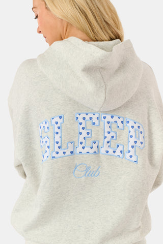 A person is wearing the Roller Rabbit Sleep Club Mimi Sweatpants in light gray, featuring wide legs and crafted from velvety soft Dream Fleece with “Sleep Club” printed in light blue on the left thigh, paired with white sneakers and a cropped white top. The background is plain white.
