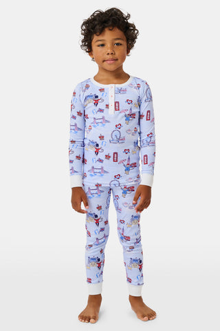 A young child with curly hair stands barefoot against a plain white background, wearing Roller Rabbit’s Kids London Monkey Pajamas—light blue long-sleeve pajamas adorned with playful, colorful patterns featuring guards, clocks, and iconic British landmarks.