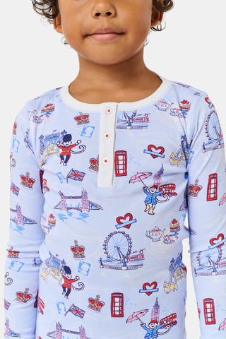 A young child with curly hair stands barefoot against a plain white background, wearing Roller Rabbit’s Kids London Monkey Pajamas—light blue long-sleeve pajamas adorned with playful, colorful patterns featuring guards, clocks, and iconic British landmarks.