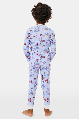 A young child with curly hair stands barefoot against a plain white background, wearing Roller Rabbit’s Kids London Monkey Pajamas—light blue long-sleeve pajamas adorned with playful, colorful patterns featuring guards, clocks, and iconic British landmarks.