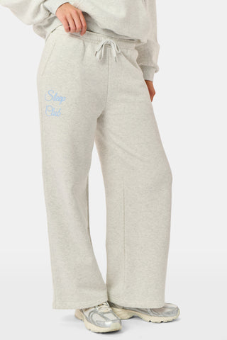 A person is wearing the Roller Rabbit Sleep Club Mimi Sweatpants in light gray, featuring wide legs and crafted from velvety soft Dream Fleece with “Sleep Club” printed in light blue on the left thigh, paired with white sneakers and a cropped white top. The background is plain white.