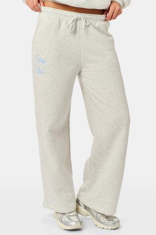 A person is wearing the Roller Rabbit Sleep Club Mimi Sweatpants in light gray, featuring wide legs and crafted from velvety soft Dream Fleece with “Sleep Club” printed in light blue on the left thigh, paired with white sneakers and a cropped white top. The background is plain white.