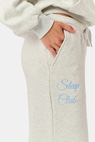 A person is wearing the Roller Rabbit Sleep Club Mimi Sweatpants in light gray, featuring wide legs and crafted from velvety soft Dream Fleece with “Sleep Club” printed in light blue on the left thigh, paired with white sneakers and a cropped white top. The background is plain white.