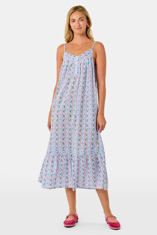 A woman wears a sleeveless SANSKRITI Mistletoe Stripe Augustine Dress in cotton with vertical floral patterns and a ruffled hem, paired with pink flat shoes.