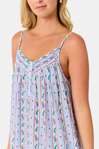 A woman wears a sleeveless SANSKRITI Mistletoe Stripe Augustine Dress in cotton with vertical floral patterns and a ruffled hem, paired with pink flat shoes.