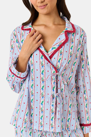 A person wearing a Mistletoe Stripe Josie Loungewear set with red trim, a matching sleep mask, and dark socks, sitting on a mossy tree trunk.