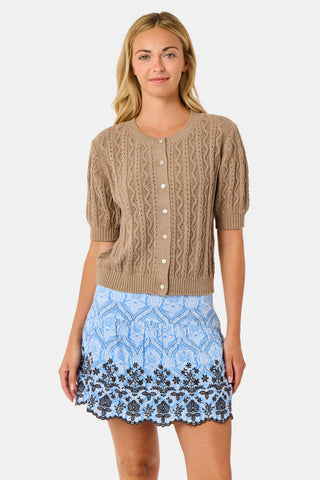A woman wears the Roller Rabbit Zig Zag Pointelle Nyla Cardigan, a short-sleeved, walnut brown button-front sweater with circular patterns, paired with a light blue patterned skirt.