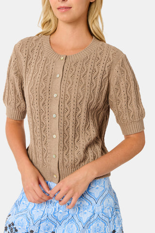A woman wears the Roller Rabbit Zig Zag Pointelle Nyla Cardigan, a short-sleeved, walnut brown button-front sweater with circular patterns, paired with a light blue patterned skirt.