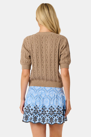 A woman wears the Roller Rabbit Zig Zag Pointelle Nyla Cardigan, a short-sleeved, walnut brown button-front sweater with circular patterns, paired with a light blue patterned skirt.