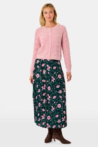 A woman wearing an AFFINA Wave Knit Moxie Cardigan in light pink over a dark green pleated skirt with a pink floral print.
