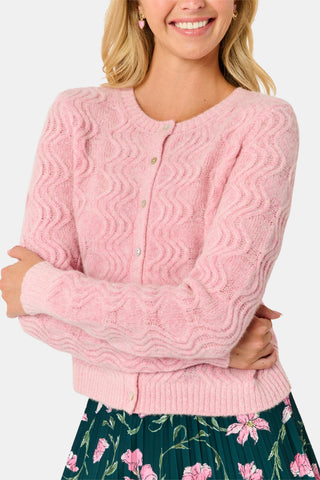 A woman wearing an AFFINA Wave Knit Moxie Cardigan in light pink over a dark green pleated skirt with a pink floral print.