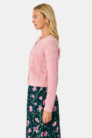 A woman wearing an AFFINA Wave Knit Moxie Cardigan in light pink over a dark green pleated skirt with a pink floral print.