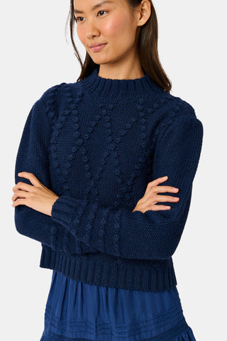 A person wearing a navy blue REVOLUTION KNITS Popcorn Knit Tessa Sweater featuring a textured, popcorn-stitch diamond pattern and a mock-neck.