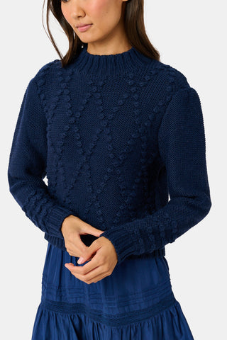 A person wearing a navy blue REVOLUTION KNITS Popcorn Knit Tessa Sweater featuring a textured, popcorn-stitch diamond pattern and a mock-neck.