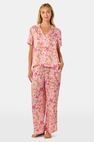 A woman stands barefoot against a plain white background wearing the Priya Floral Alvie Set by Roller Rabbit—a matching pink pajama set featuring a floral pattern, short-sleeve button-up top, and loose pants crafted from washable silk loungewear.