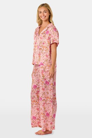 A woman stands barefoot against a plain white background wearing the Priya Floral Alvie Set by Roller Rabbit—a matching pink pajama set featuring a floral pattern, short-sleeve button-up top, and loose pants crafted from washable silk loungewear.