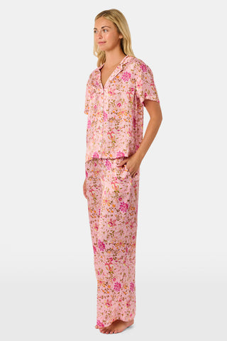 A woman stands barefoot against a plain white background wearing the Priya Floral Alvie Set by Roller Rabbit—a matching pink pajama set featuring a floral pattern, short-sleeve button-up top, and loose pants crafted from washable silk loungewear.