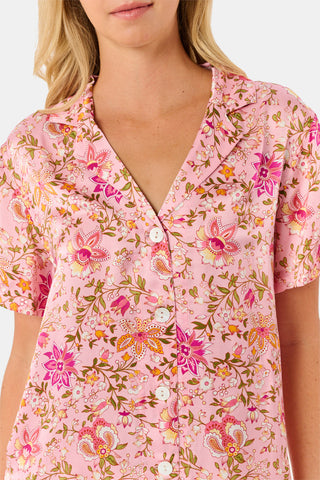 A woman stands barefoot against a plain white background wearing the Priya Floral Alvie Set by Roller Rabbit—a matching pink pajama set featuring a floral pattern, short-sleeve button-up top, and loose pants crafted from washable silk loungewear.