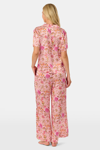 A woman stands barefoot against a plain white background wearing the Priya Floral Alvie Set by Roller Rabbit—a matching pink pajama set featuring a floral pattern, short-sleeve button-up top, and loose pants crafted from washable silk loungewear.