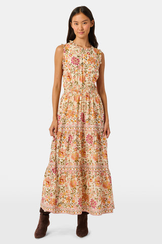 A woman in the Priya Floral Milani Dress by Roller Rabbit and boots stands on grass, holding a hat filled with red apples. Trees and sunlight fill the background.