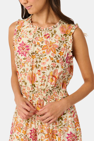 A woman in the Priya Floral Milani Dress by Roller Rabbit and boots stands on grass, holding a hat filled with red apples. Trees and sunlight fill the background.