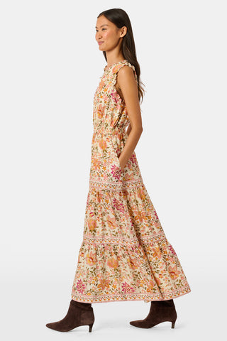 A woman in the Priya Floral Milani Dress by Roller Rabbit and boots stands on grass, holding a hat filled with red apples. Trees and sunlight fill the background.