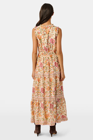 A woman in the Priya Floral Milani Dress by Roller Rabbit and boots stands on grass, holding a hat filled with red apples. Trees and sunlight fill the background.