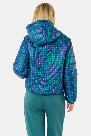A woman wearing a SOPHISTICA Plaid Hearts Reversible Puffer Jacket, a hooded, zip-front nylon puffer in an emerald green and blue plaid pattern accented by small pink hearts.