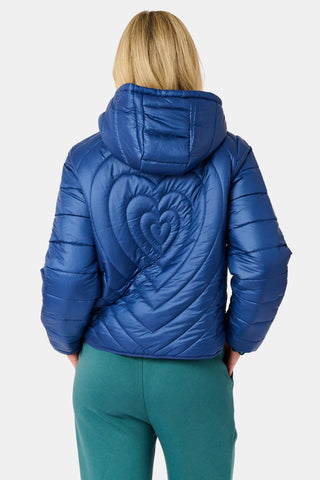 A woman wearing a SOPHISTICA Plaid Hearts Reversible Puffer Jacket, a hooded, zip-front nylon puffer in an emerald green and blue plaid pattern accented by small pink hearts.