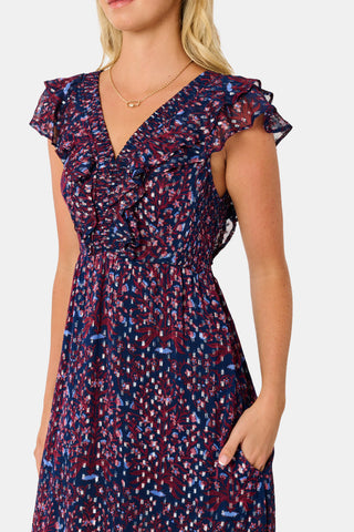 A woman wears a burgundy botanical print SOPHISTICA Rhone Ondine Dress featuring flutter sleeves, a ruffled neckline, and side pockets, paired with black platform heels for a holiday look.