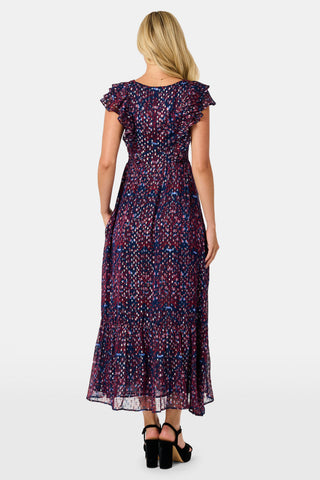 A woman wears a burgundy botanical print SOPHISTICA Rhone Ondine Dress featuring flutter sleeves, a ruffled neckline, and side pockets, paired with black platform heels for a holiday look.