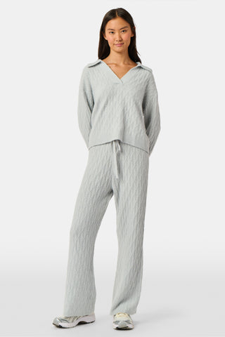 A woman wearing a light gray Cable Knots Rocio Sweater by Roller Rabbit, featuring a textured knit and V-neck, paired with matching knit pants, stands against a plain white background.
