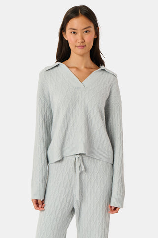 A woman wearing a light gray Cable Knots Rocio Sweater by Roller Rabbit, featuring a textured knit and V-neck, paired with matching knit pants, stands against a plain white background.