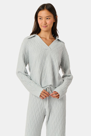 A woman wearing a light gray Cable Knots Rocio Sweater by Roller Rabbit, featuring a textured knit and V-neck, paired with matching knit pants, stands against a plain white background.