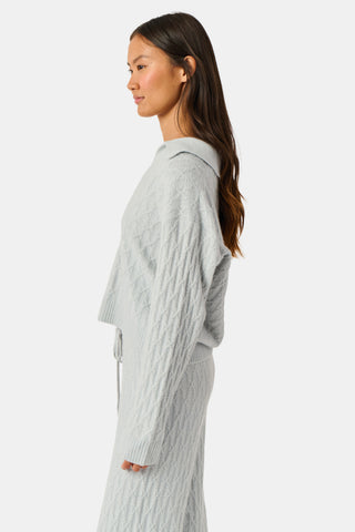 A woman wearing a light gray Cable Knots Rocio Sweater by Roller Rabbit, featuring a textured knit and V-neck, paired with matching knit pants, stands against a plain white background.