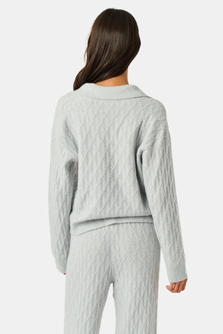 A woman wearing a light gray Cable Knots Rocio Sweater by Roller Rabbit, featuring a textured knit and V-neck, paired with matching knit pants, stands against a plain white background.